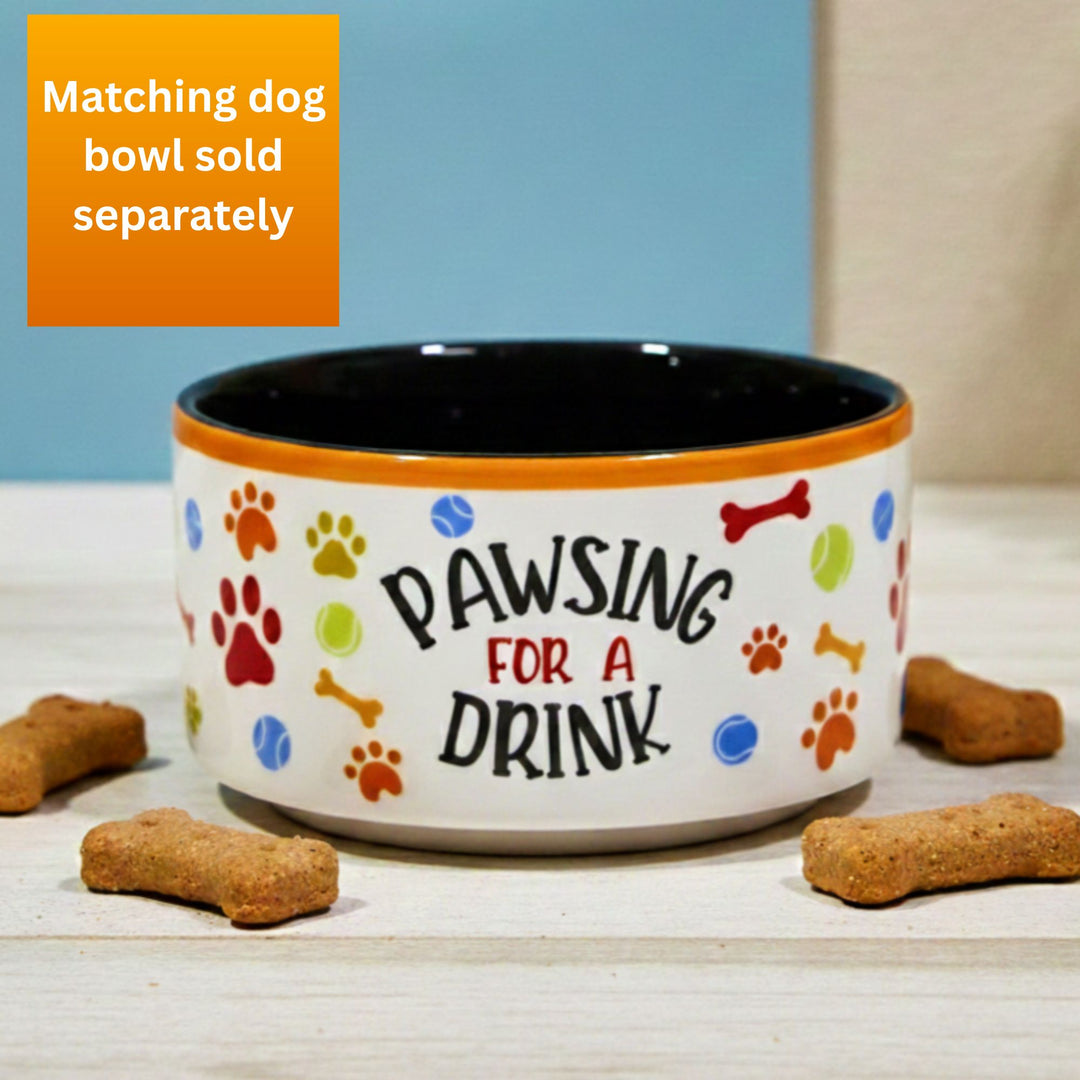 Dog bowl with 'Pawsing for a Drink' text and colorful paw print design on a wooden surface with dog treats. Sold separately from a matching mug.