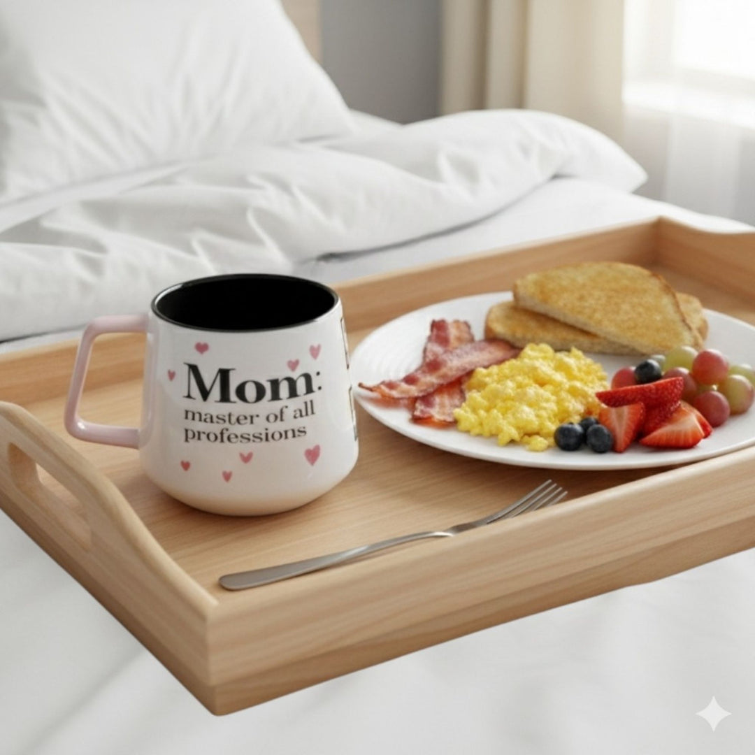 Lifestyle image of a mug with text on it saying 'Mom: master of all professions',   next to a plate of breakfast food on a tray.